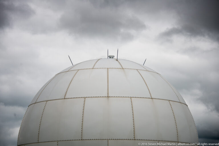 A close view of a white spherical dome shows curved panel seams, fasteners, and small fittings against a cloudy sky.