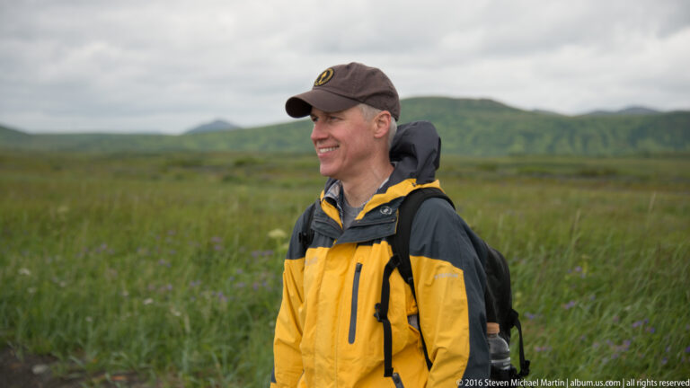 A visitor in a yellow-and-black jacket and backpack stands among tundra vegetation with low hills behind him.