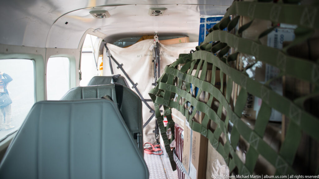 Gray aircraft seats line one side of a narrow cabin while secured packs and equipment occupy the other.