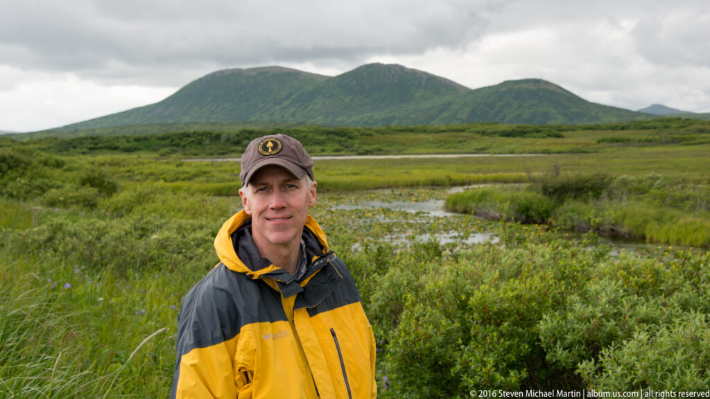 A visitor in a gray-and-yellow jacket stands before a wetland and a rounded green mountain.