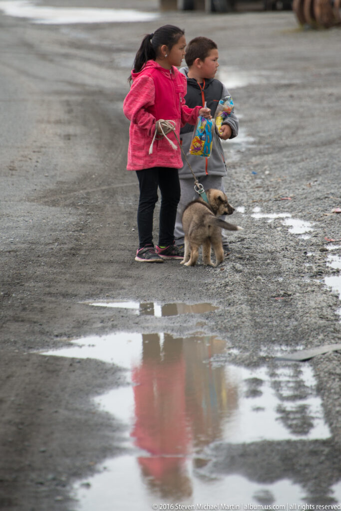 Two children in bright jackets stand with a small dog beside puddles on a muddy road.