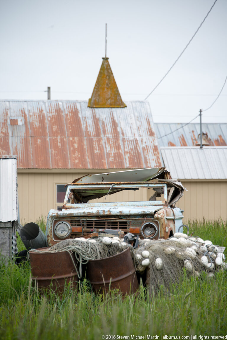 A rusted pickup sits behind nets, tubs, and other equipment in a grassy yard beside metal-roofed buildings.