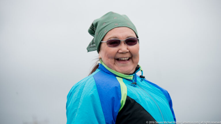 A smiling woman in a green headscarf and tinted sunglasses wears a bright blue jacket.