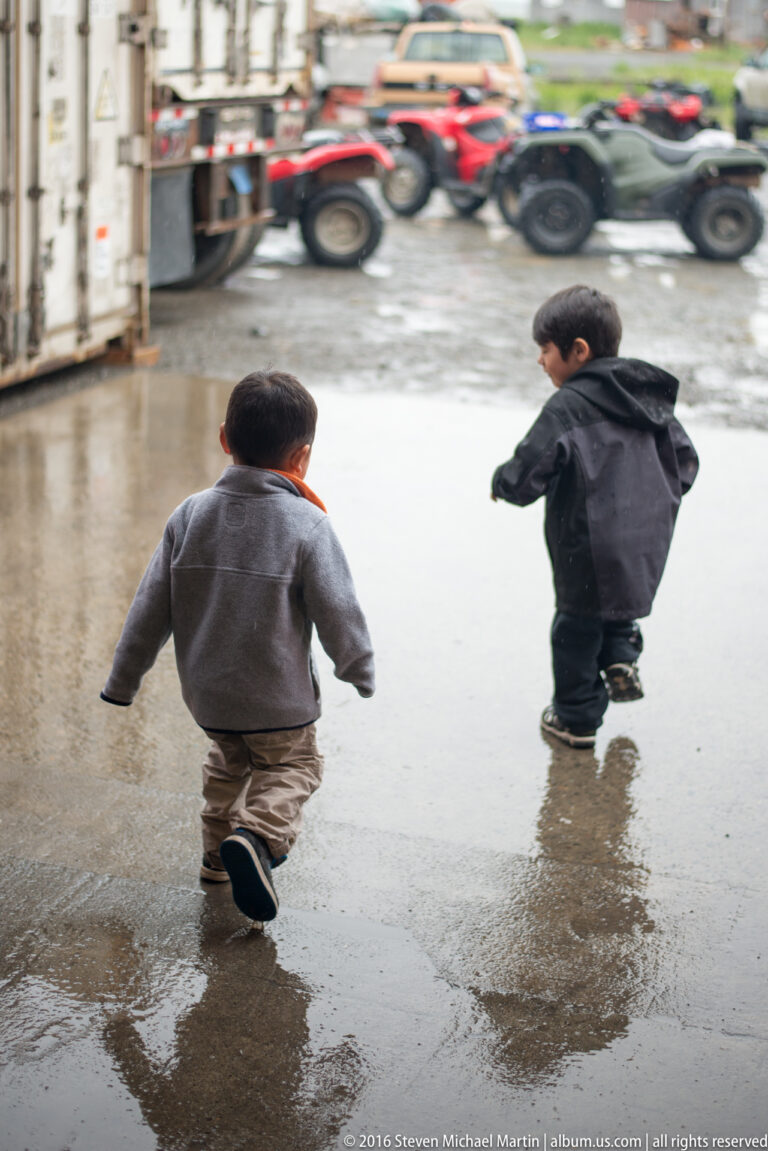 Two children walk through a puddle on a muddy street, with parked ATVs and buildings behind them.