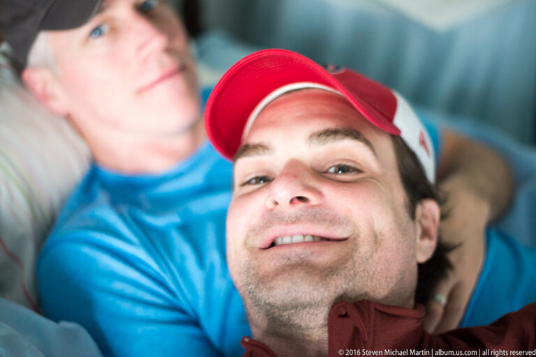 A smiling man in a red-and-white cap is seen close up, with another person blurred behind him.