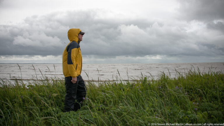 A man in a yellow hooded jacket stands in coastal grass and looks across the water beneath dark clouds.