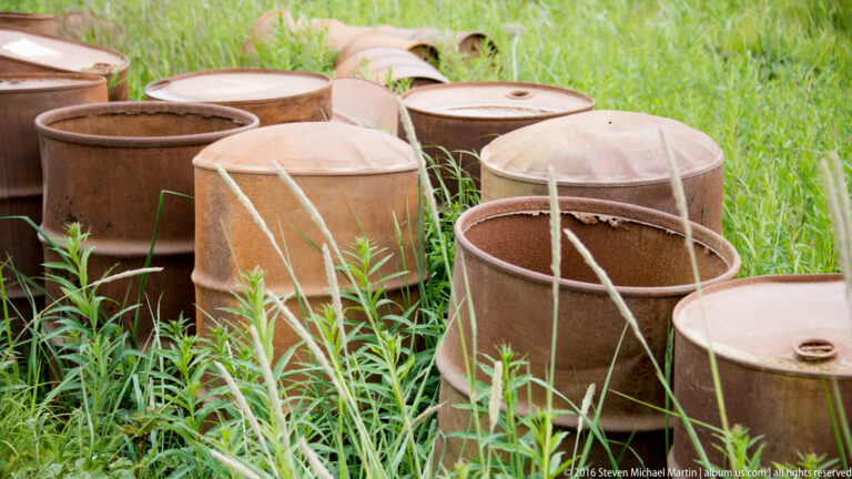 Rusty open metal cylinders with crossbars sit among tall green grass and weeds.
