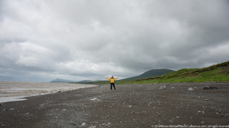 A person in a yellow jacket stands with arms extended on a broad beach beneath heavy clouds.