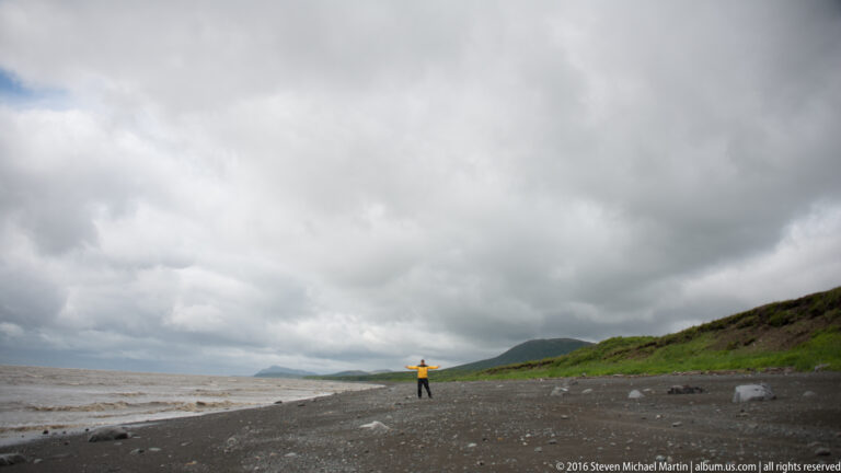 A distant person in yellow stands on a long beach beneath clouds, with a green headland at right.