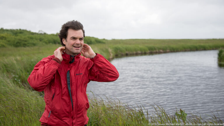 A man in a red jacket adjusts his collar beside a narrow channel bordered by tall green grasses.