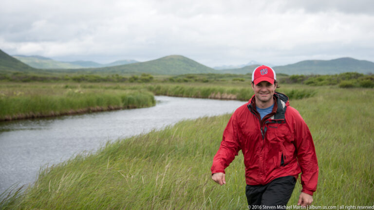 A man in a red jacket and cap walks through tall grass beside a winding channel, with hills beyond.