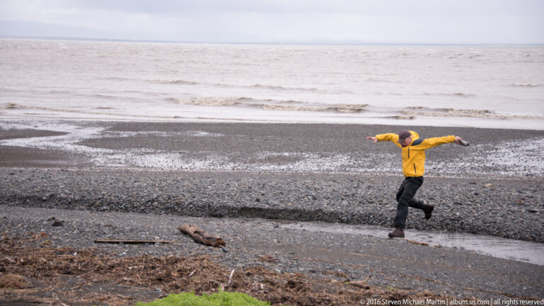 A person in a yellow jacket steps across a shallow tidal channel with arms outstretched.