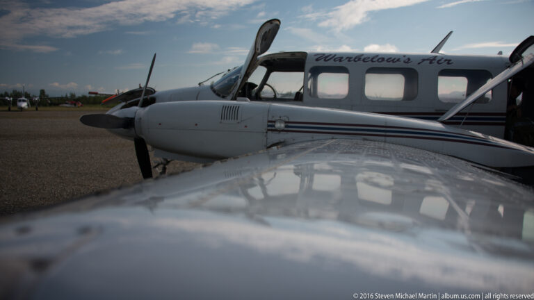 A Warbelow’s Air plane sits on an airstrip, its reflective wing filling the foreground beneath the cabin windows.