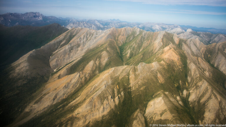 Pale folded slopes with dark gullies and sparse green vegetation descend into a shadowed basin.