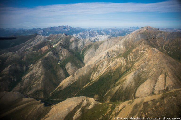 Pale angular ridges and dark valleys repeat across the landscape, with green drainage corridors below a distant skyline.