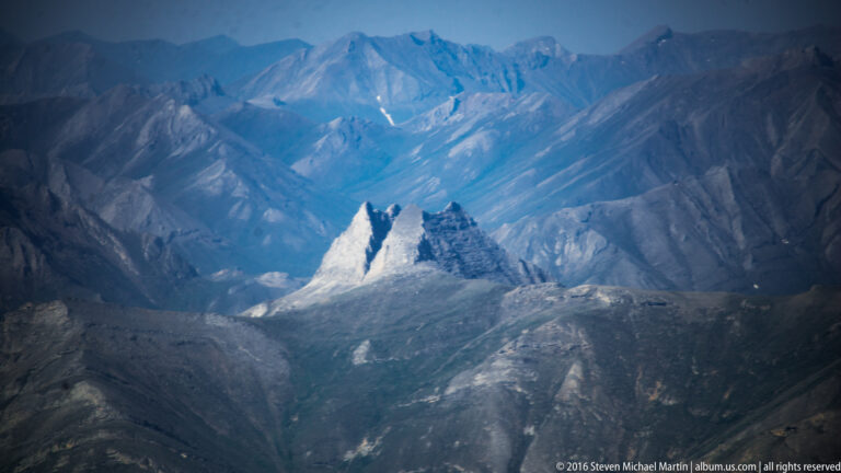 Two pale pointed rock spires rise from a shadowed mountain basin, backed by hazy ranges.