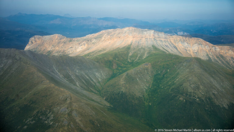 A rounded pale summit is lit above darker tundra-covered slopes and hazy distant ridges.