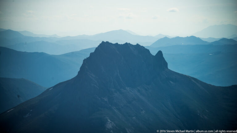 A jagged dark summit stands above a nearer ridge against hazy blue mountain layers.