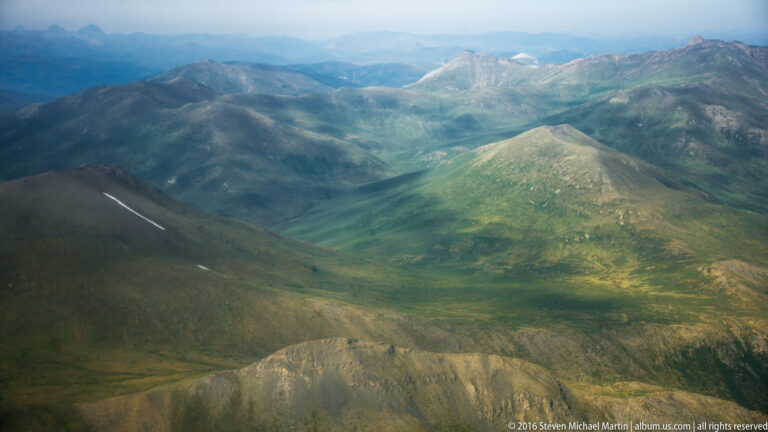 Rounded tundra ridges descend toward a green valley and distant mountains, with a few snow patches on the left.