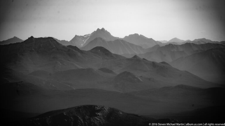 A pointed peak rises above layered dark mountain ridges fading into a pale sky in a monochrome aerial view.
