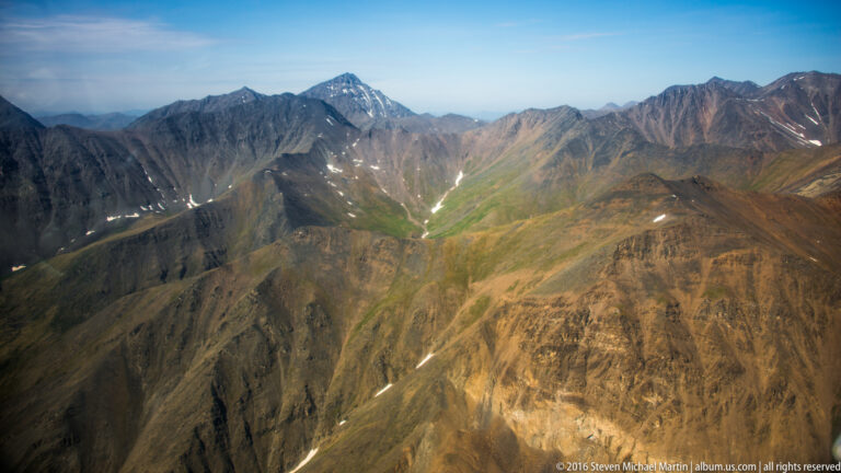 Green drainage lines descend through a mountain basin bordered by dark ridges with scattered snow patches.