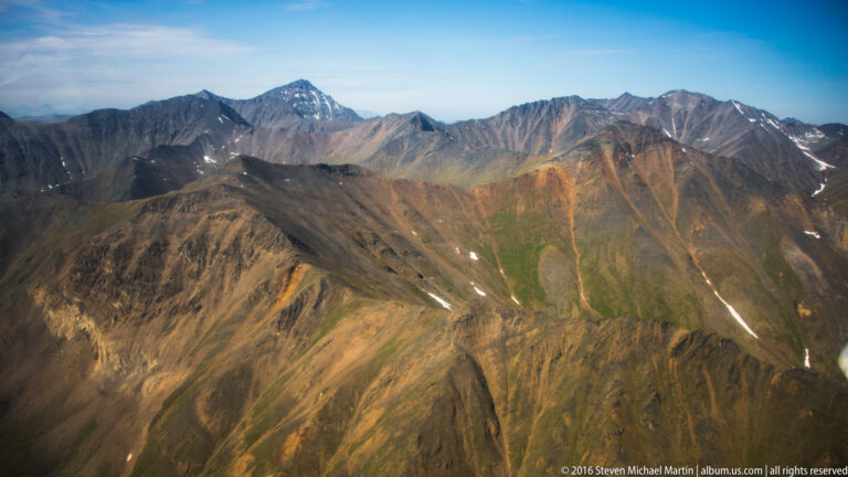 A dark foreground ridge leads toward a pale distant summit beyond several brown mountain ranges.