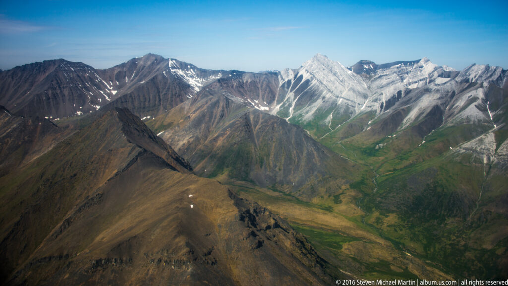 Brown foreground ridges frame a green valley leading toward a pale, snow-streaked mountain massif.