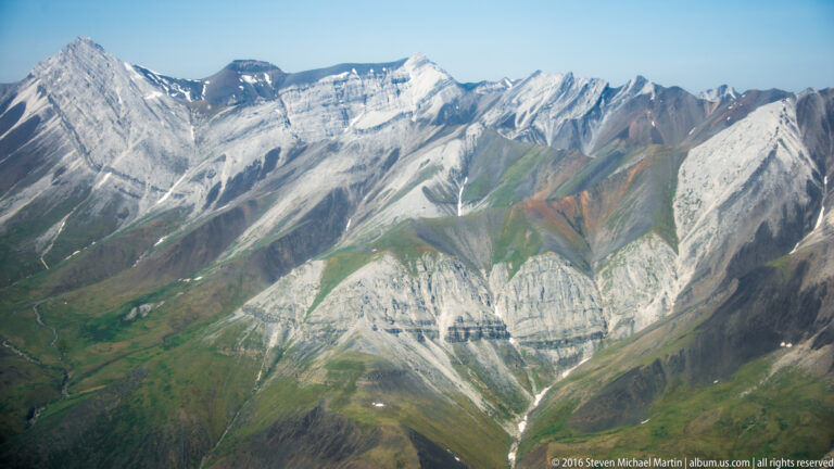 A light-colored mountain face rises above a green valley, framed by darker peaks and streaked with snow in shaded ravines.