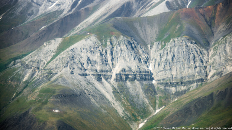 A pale mountain wall with vertical erosion channels and dark horizontal bands rises above green slopes.
