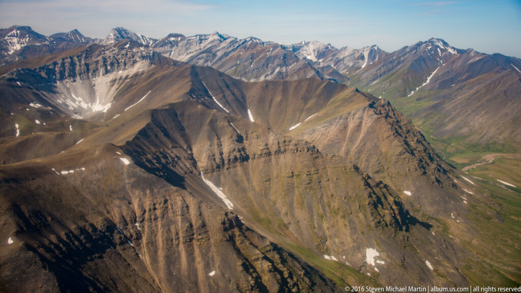 Intersecting brown and gray mountain ridges descend toward a green valley with a pale streambed at right.