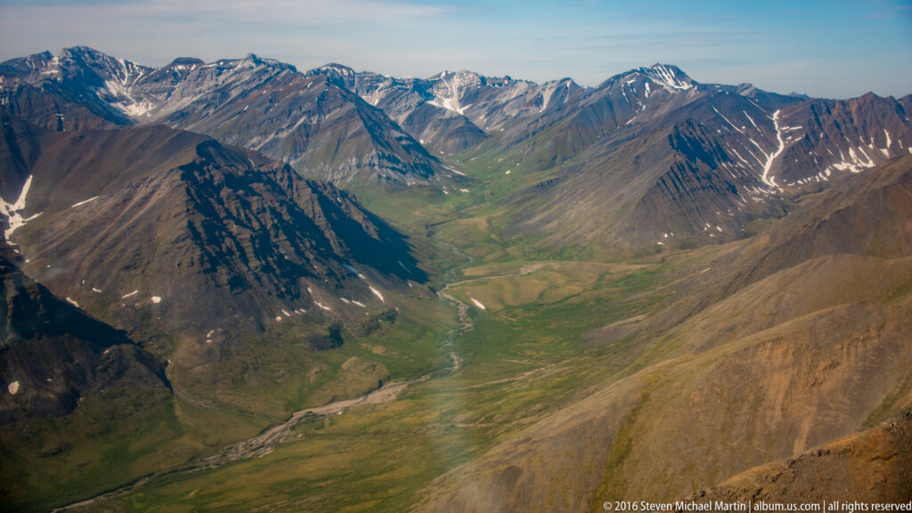 A broad green valley and pale riverbed extend between jagged, snow-streaked mountain ranges toward the horizon.