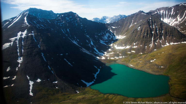 A turquoise lake lies below converging mountain ridges, with snow patches on sunlit slopes and deep shadow along the left wall.