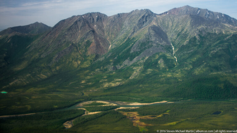 Aerial view of a steep mountain valley with pale drainage lines and a braided river crossing its green floor.