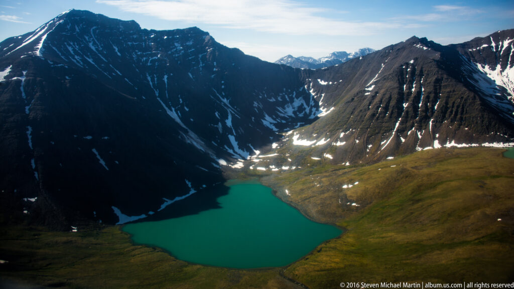A turquoise lake fills the bottom of a mountain basin, ringed by dark slopes, snow-filled ravines, and tundra.