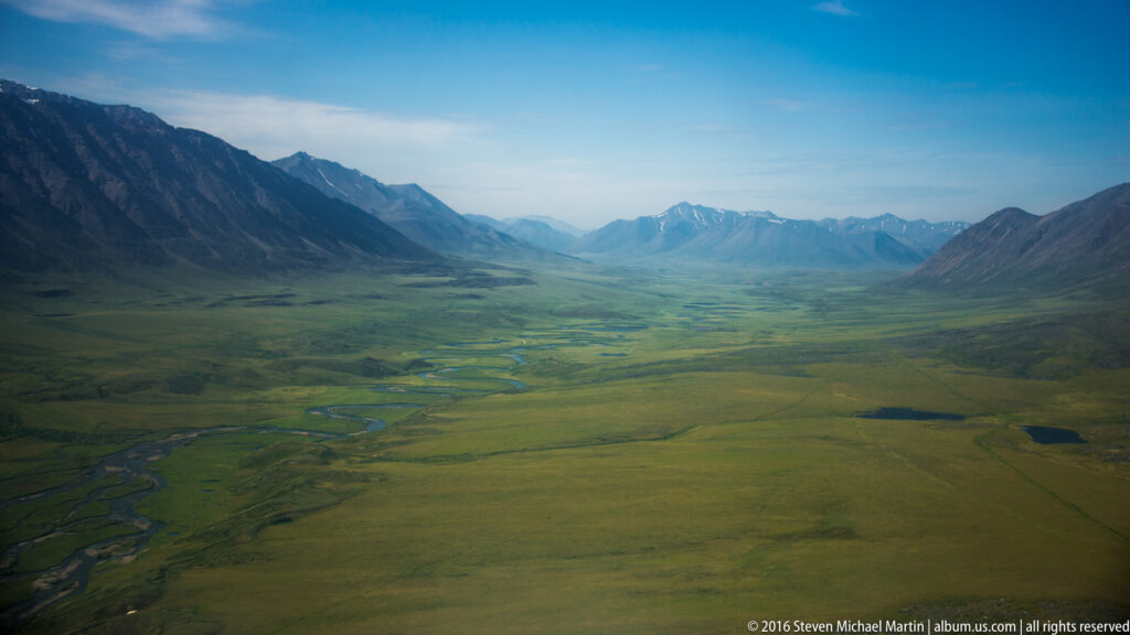 A braided river crosses a broad tundra valley between distant mountain ranges, with small ponds scattered across the plain.