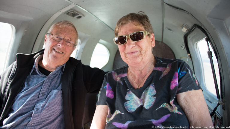 Two older passengers smile at the camera from adjacent seats inside a small aircraft cabin.