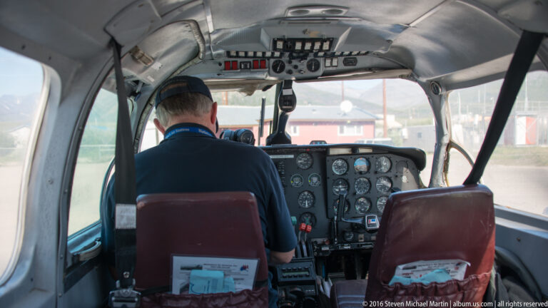 A pilot sits at the controls of a small aircraft, surrounded by analog instruments and looking through the windshield toward buildings.