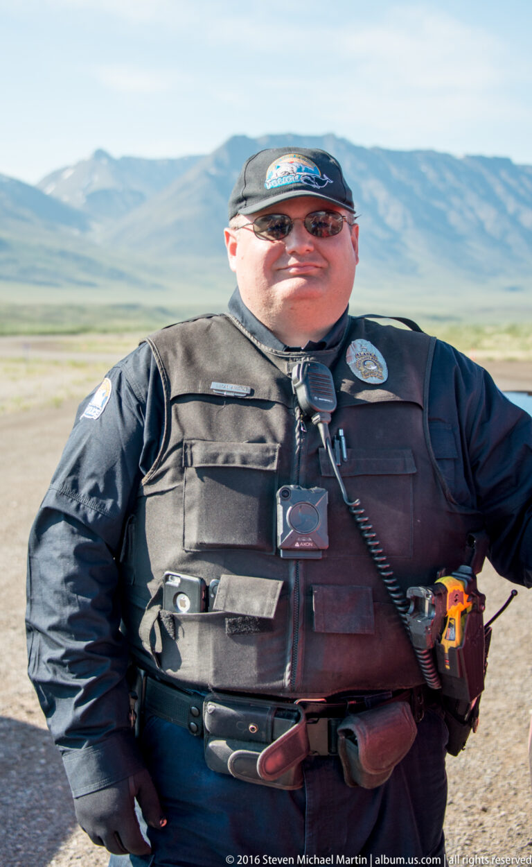 A man wearing a dark equipment vest, radio, cap, and sunglasses stands before open tundra.