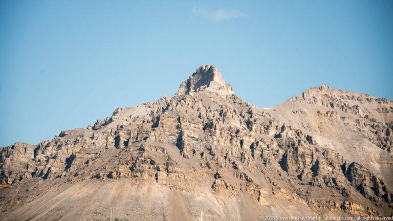 A sharply pointed pale mountain summit rises above layered, gullied rock slopes.