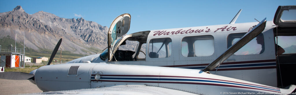 White Warbelow’s Air bush plane parked at a small airfield with mountains behind it.