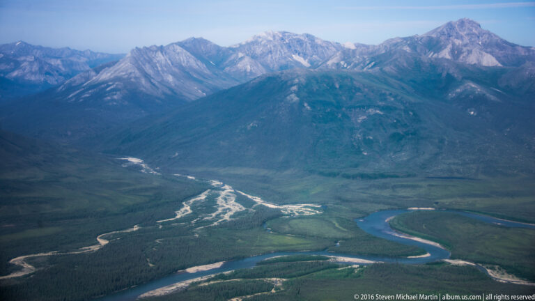 A winding river loops across a forested valley and divides into braided channels beneath steep mountains.