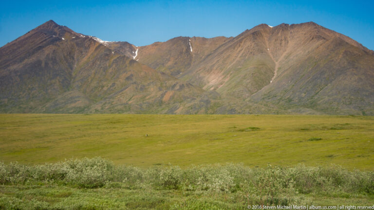Open tundra with low shrubs stretches below a mountain ridge with a small snow remnant.