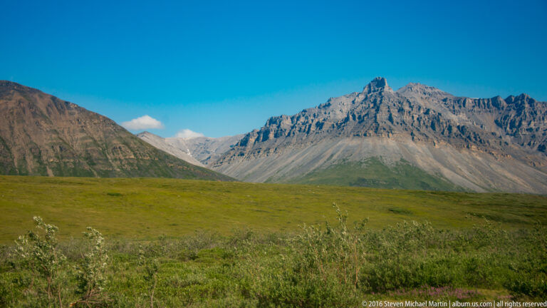 Low tundra vegetation leads toward a rugged mountain ridge under clear blue sky.