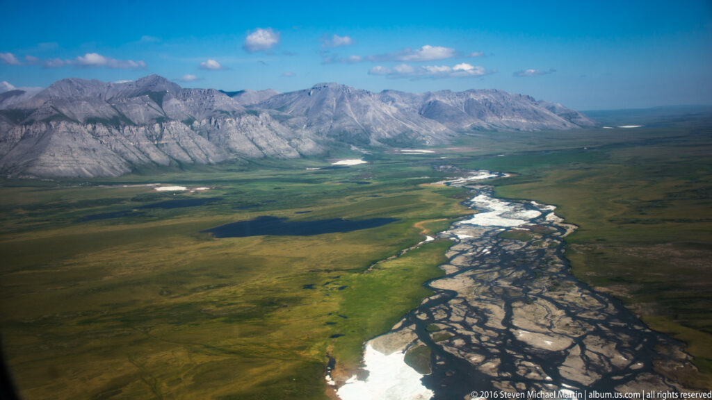 A braided river cuts diagonally across a green lowland toward distant mountains.