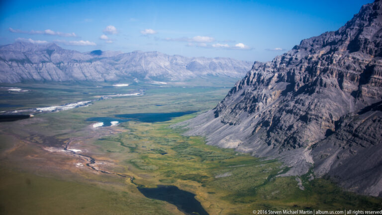 A massive rocky mountain wall borders a green valley with small lakes and a winding stream.