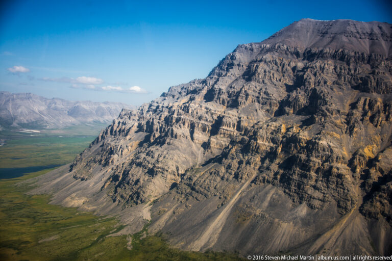 A rugged layered escarpment rises above a broad green plain, with distant mountains beyond.