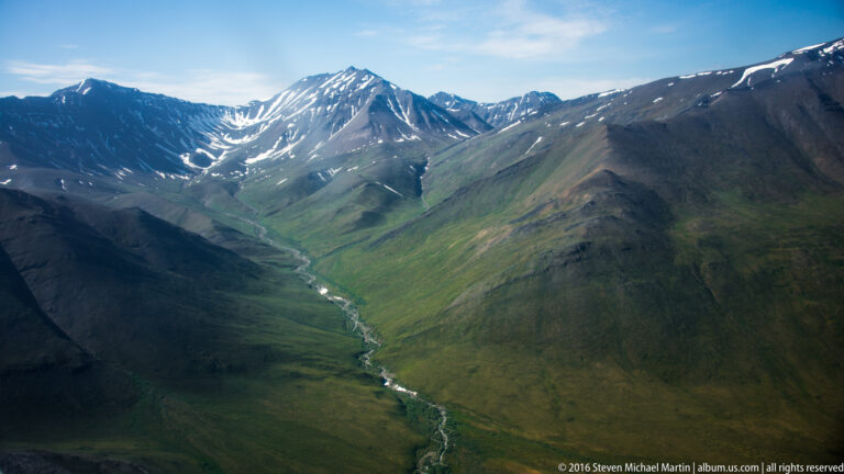 A thin stream runs down a green valley toward a snow-streaked ridge between dark mountains.