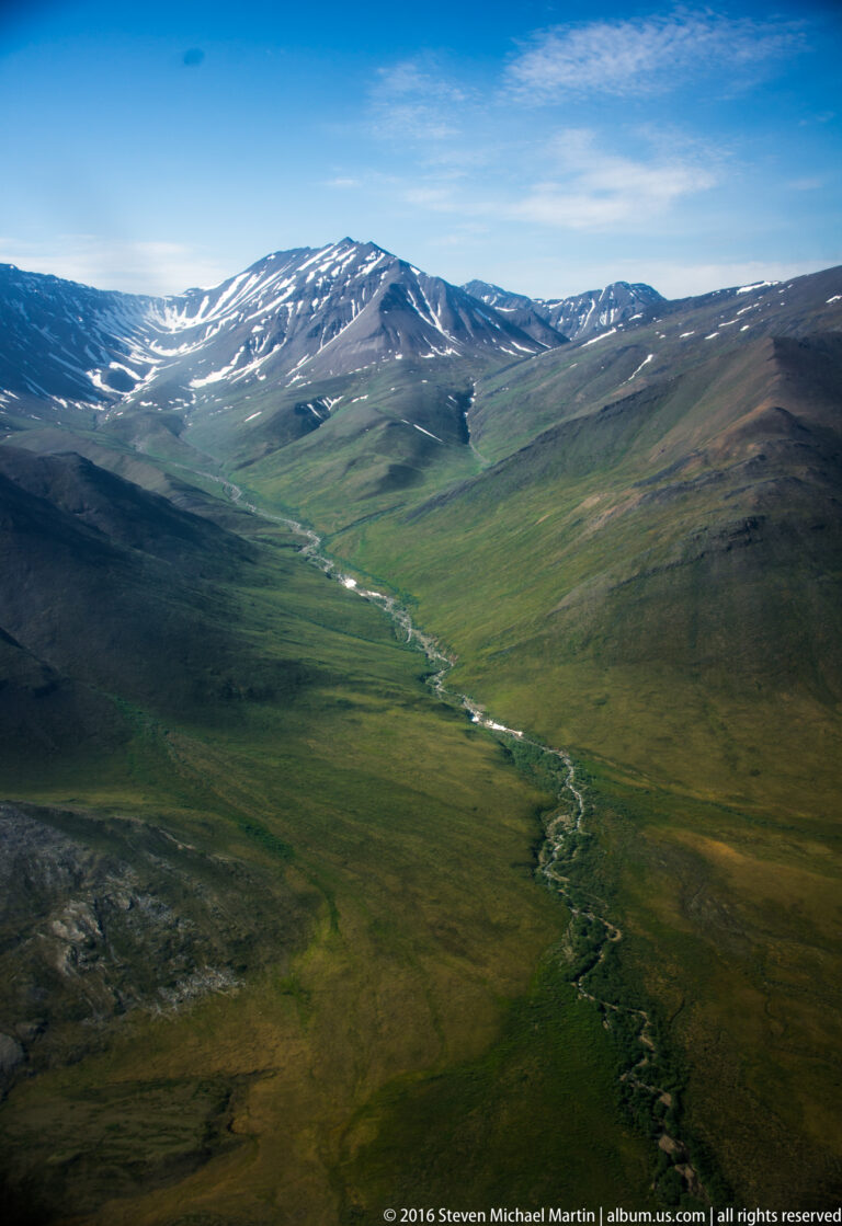 A winding stream crosses a broad green valley toward snow-streaked distant ridges.