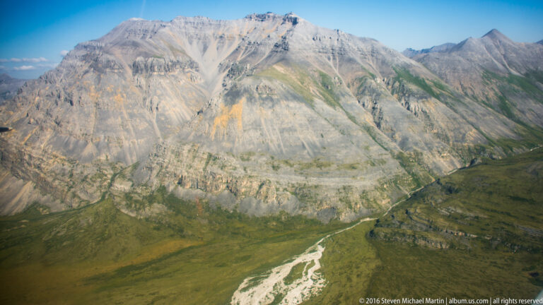 A steep pale mountain wall descends toward a braided stream and distant valley.