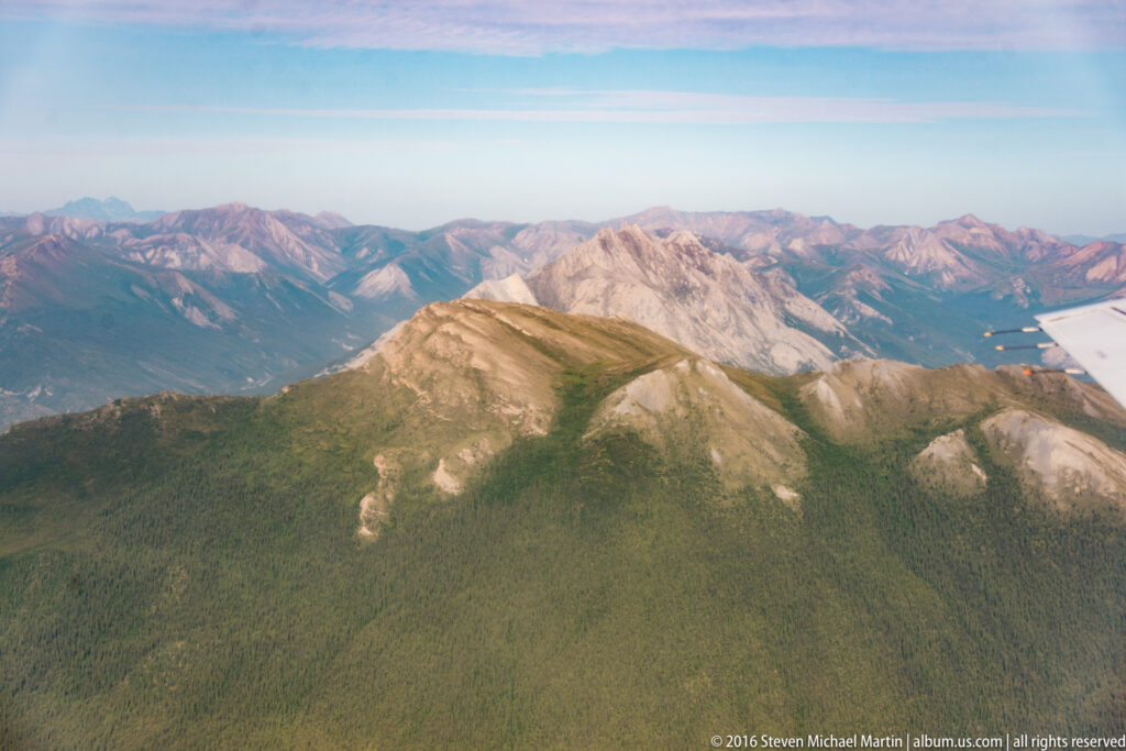 A forested ridge and pale rocky mountains recede beneath a clear sky, with part of an aircraft visible at the right edge.